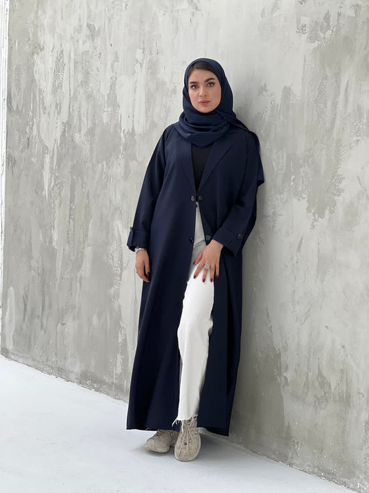 A woman wearing a Navy blue coat-style abaya with a Sheila headscarf, standing against a gray textured background.