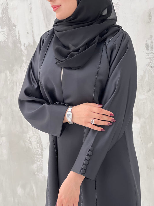 A woman wearing a black Satin abaya with handwork, standing against a grey background.
