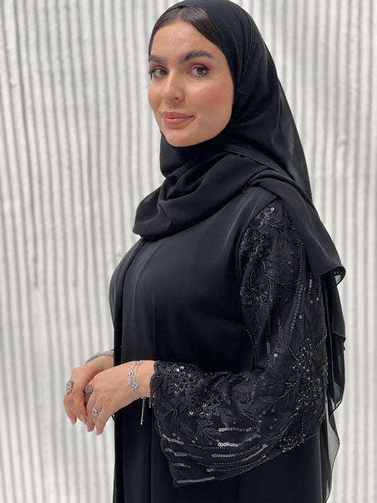 A woman wearing a Black Abaya with sequin-embroidered sleeves, with a matching Sheila standing against a white background.