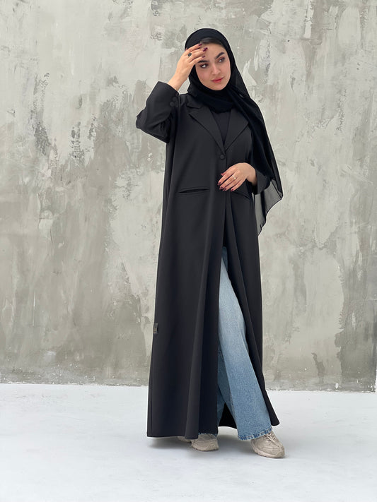 A woman wearing a Classic Black abaya with a black sheila, over a black outfit, with blue jeans and beige shoes, standing against a gray background.