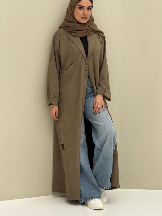 A woman wearing a khaki shamwa Coat style abaya with lapel collar, Button accents on front, angled pockets and matching sheila on a cream background