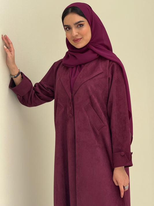 A woman wearing a maroon shamwa Coat style abaya with lapel collar, Button accents on front, angled pockets and matching sheila on a cream background
