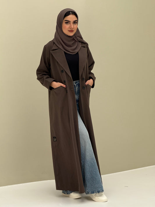 A woman wearing a dark brown abaya with double-breasted buttons, buttoned cuffs, a chest pocket, and a matching sheila paired with blue jeans and white shoes.