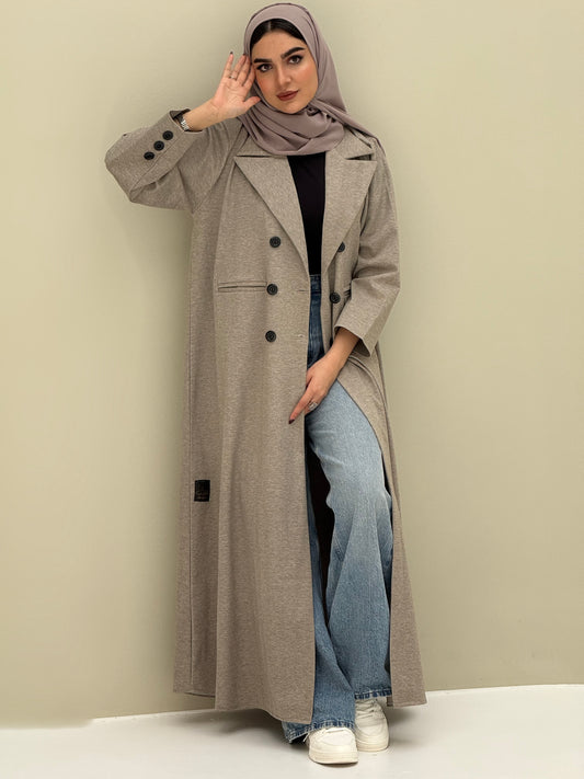 A woman wearing a light brown abaya with double-breasted buttons, buttoned cuffs, a chest pocket, and a matching sheila paired with blue jeans and white shoes.