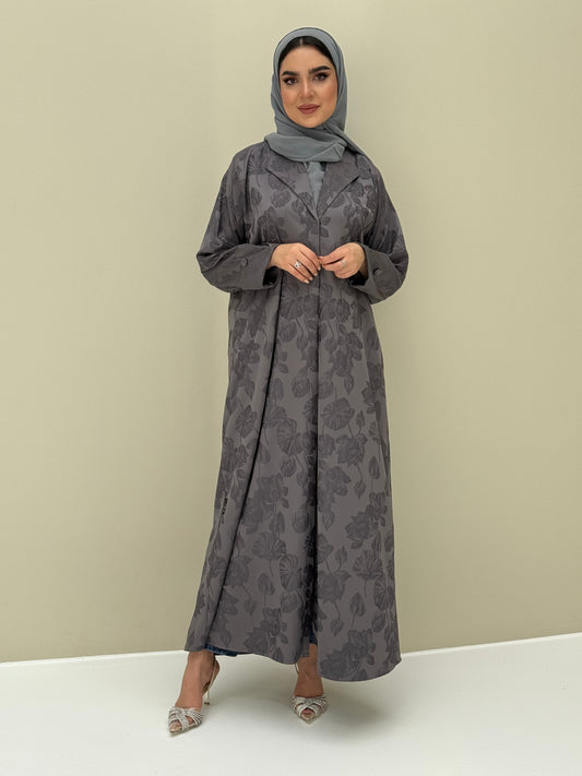 A woman wearing a steel grey abaya with a jacquard fabric, including a matching sheila headscarf, standing against a beige background.