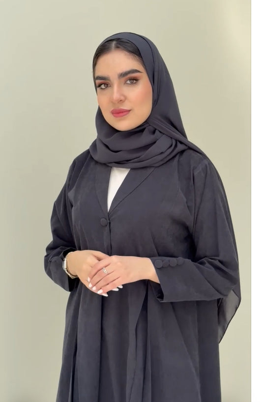 A woman standing in a neutral pose wearing a dark grey Ice Cool Abaya flared coat style with lapel collar and a matching sheila, white shoes.