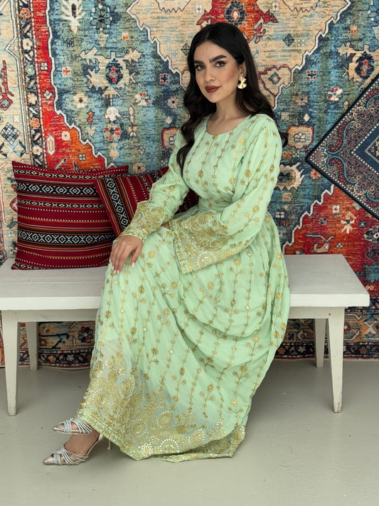 A woman seated on a bench against a colorful, patterned backdrop, wearing a mint green A-line Jalabiya with wide sleeves with gold embroidered details and long sleeves.