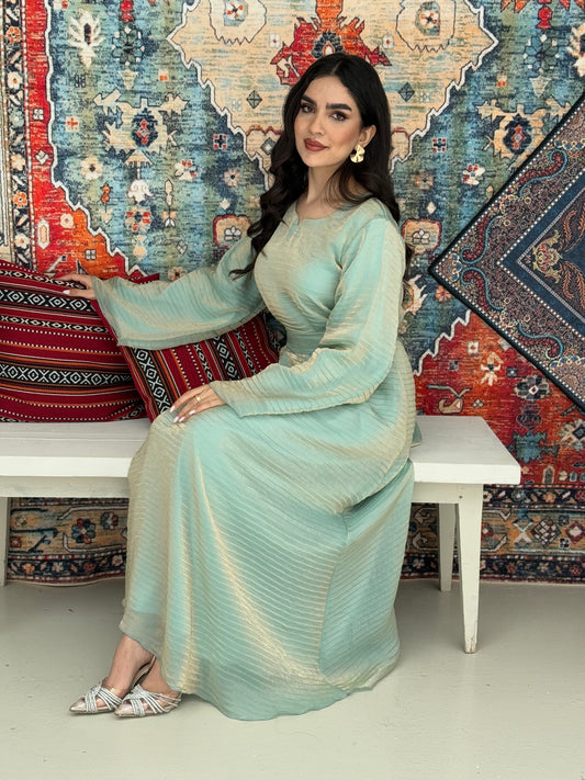 A woman standing in front of a decorative rug, wearing a mint gold Shimmer Organza dress with long sleeves and a belt, with her hair styled in an updo.