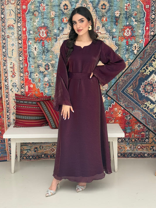 A woman standing in front of a decorative rug, wearing a purple Shimmer Organza dress with long sleeves and a belt, with her hair styled in an updo.