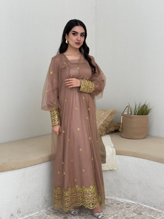 A woman standing in a tropical setting, wearing a mocha brown jalabiya dress with gold embroidery and a belt, posing with a smile.