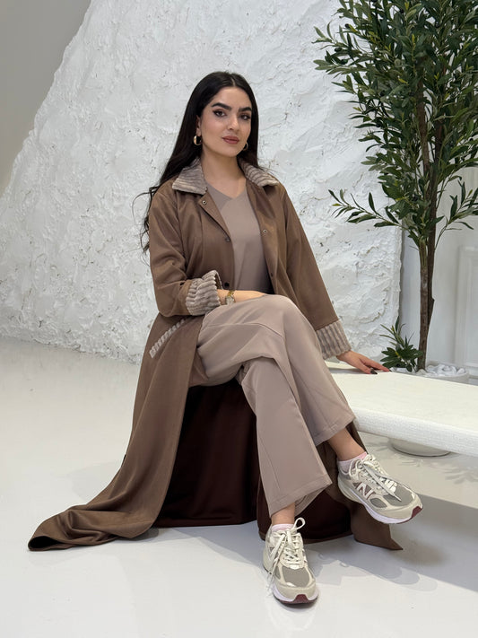 Woman in a Light Brown Shamwa Abaya and beige pants sitting on a textured white floor with green plants.