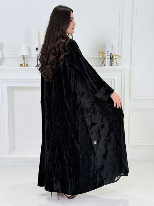 woman wearing a dark black plissé velvet abaya coat standing in front of a white door with gold handles.