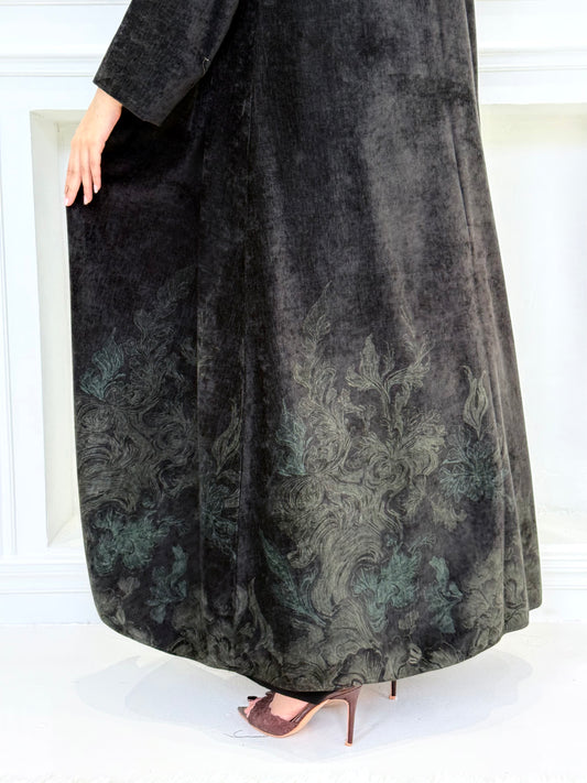 Woman in a long Black Velvet Abaya with Forest Green Details standing in front of a white door.