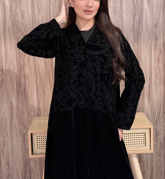 Woman wearing Premium Black Velvet Abaya with Floral Embroidery against a wooden wall.