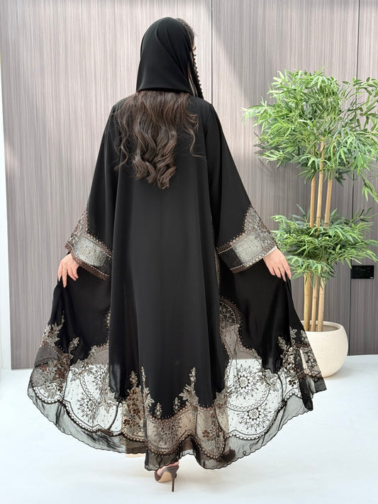 Woman wearing an open-front black abaya featuring intricate sheer panels with bronze, gold, and silver embroidery with matching sheila standing in a room with a plant and lamp.