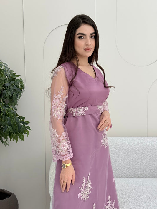 Woman in an elegant dusty purple modest jalabiya designed with a flattering V-neck, floral embroidered sheer sleeves, and a beautifully structured matching waist belt. standing against a white wall.