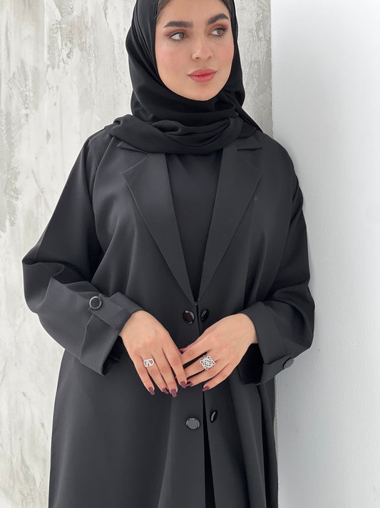 A woman wearing a black coat-style abaya with a Sheila headscarf, standing against a gray textured background.