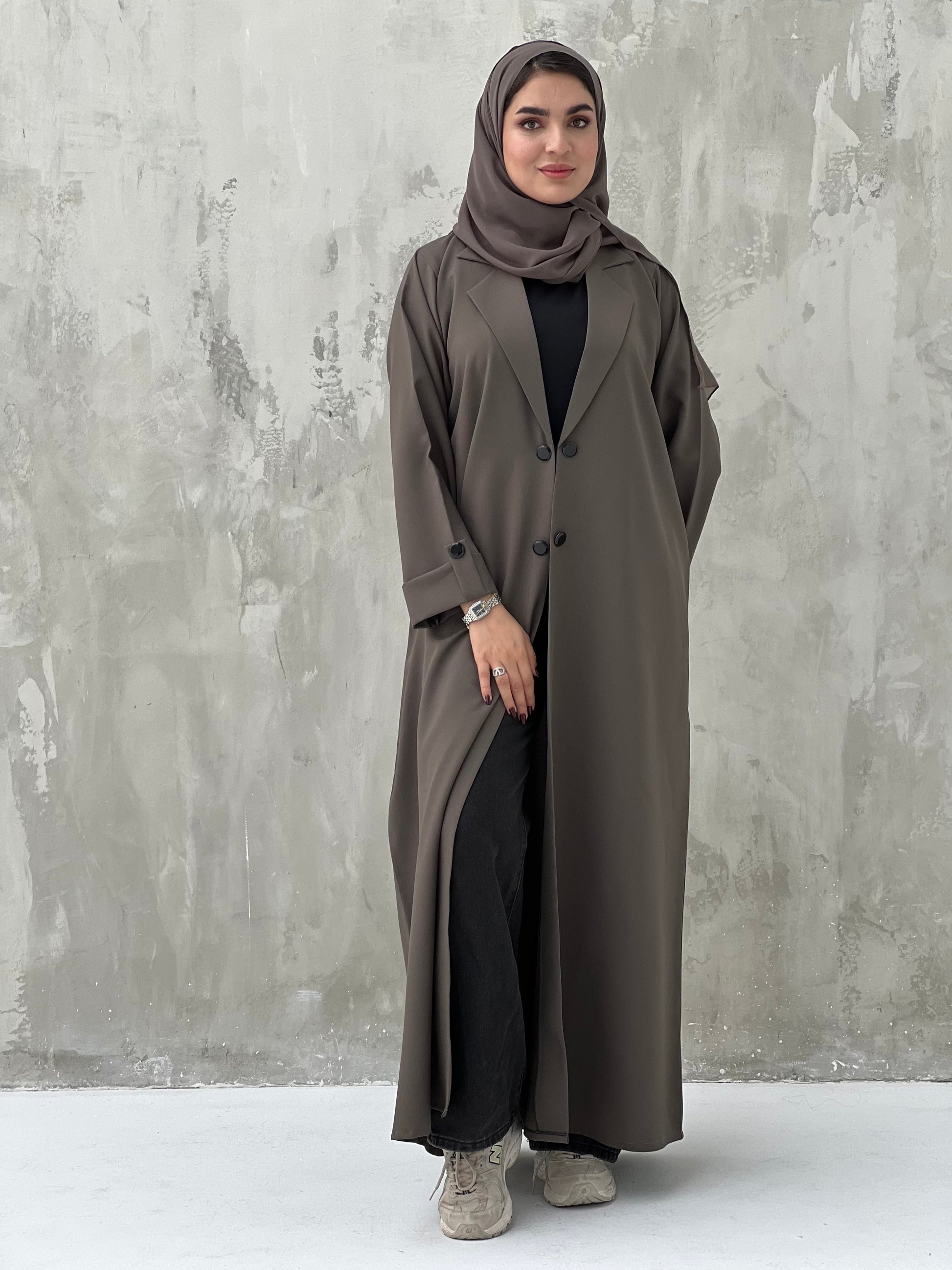 A woman wearing a Dark Brown coat-style abaya with a Sheila headscarf, standing against a gray textured background.