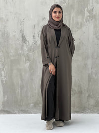 A woman wearing a Dark Brown coat-style abaya with a Sheila headscarf, standing against a gray textured background.