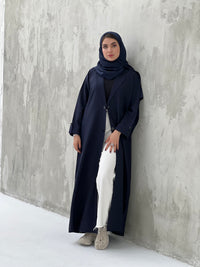 A woman wearing a Navy blue coat-style abaya with a Sheila headscarf, standing against a gray textured background.
