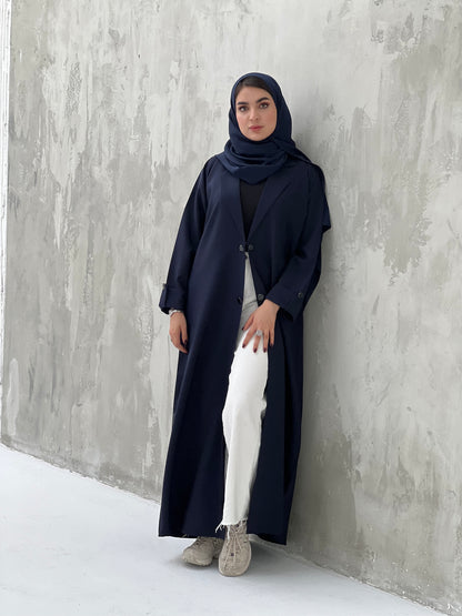 A woman wearing a Navy blue coat-style abaya with a Sheila headscarf, standing against a gray textured background.