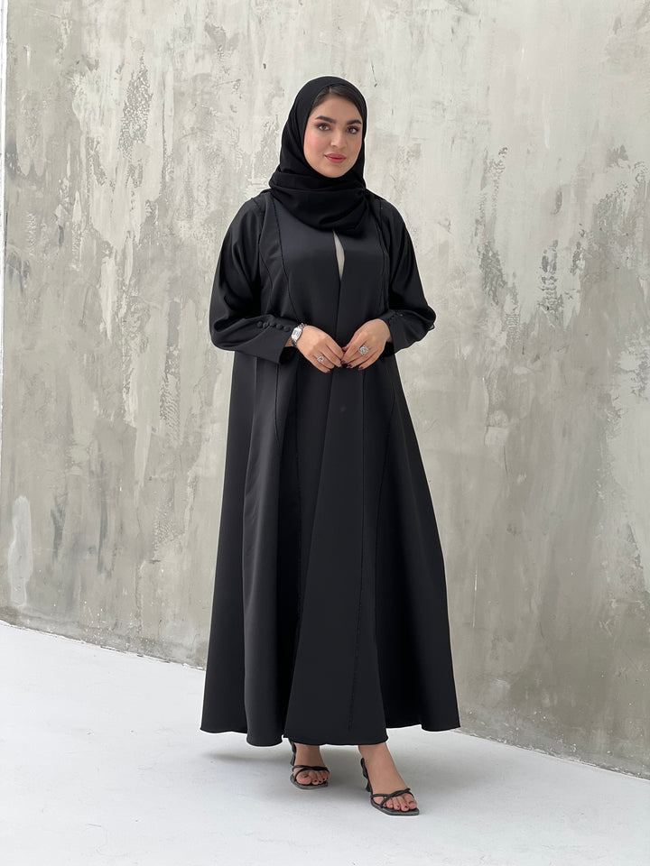 A woman wearing a black Satin abaya with handwork, standing against a grey background.