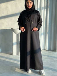 A woman wearing a Black Blazer-Style Abaya with long sleeves and a matching Sheila, standing indoors.