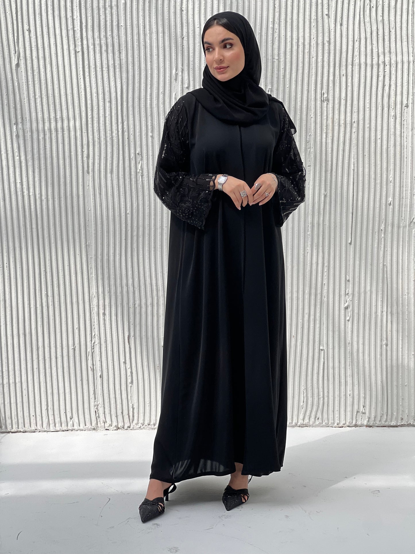 A woman wearing a Black Abaya with sequin-embroidered sleeves, with a matching Sheila standing against a white background.