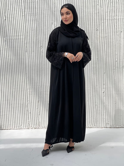 A woman wearing a Black Abaya with sequin-embroidered sleeves, with a matching Sheila standing against a white background.
