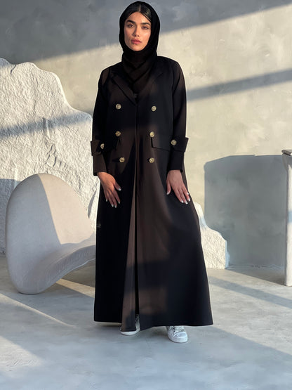 A woman wearing a Black coat-style Abaya with a lapel collar and statement gold buttons with a matching Sheila