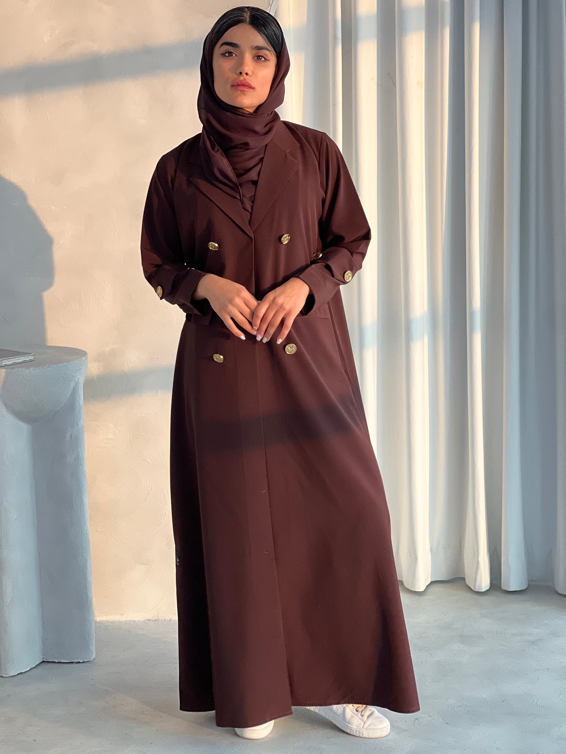 A woman wearing a Dark Brown coat-style Abaya with a lapel collar and statement gold buttons with a matching Sheila