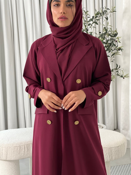 A woman wearing a Maroon coat-style Abaya with a lapel collar and statement gold buttons with a matching Sheila