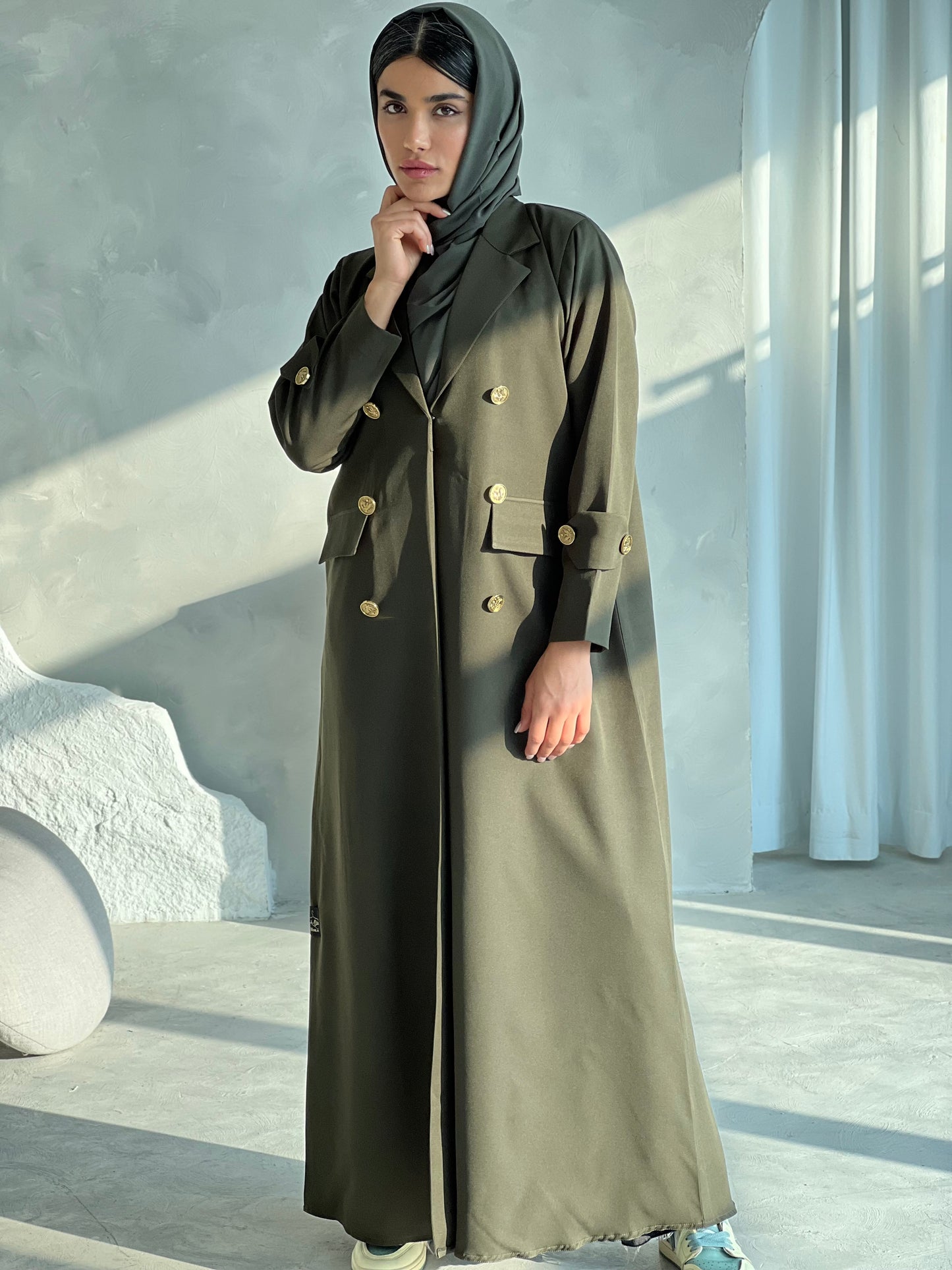 A woman wearing an Olive Green coat-style Abaya with a lapel collar and statement gold buttons with a matching Sheila