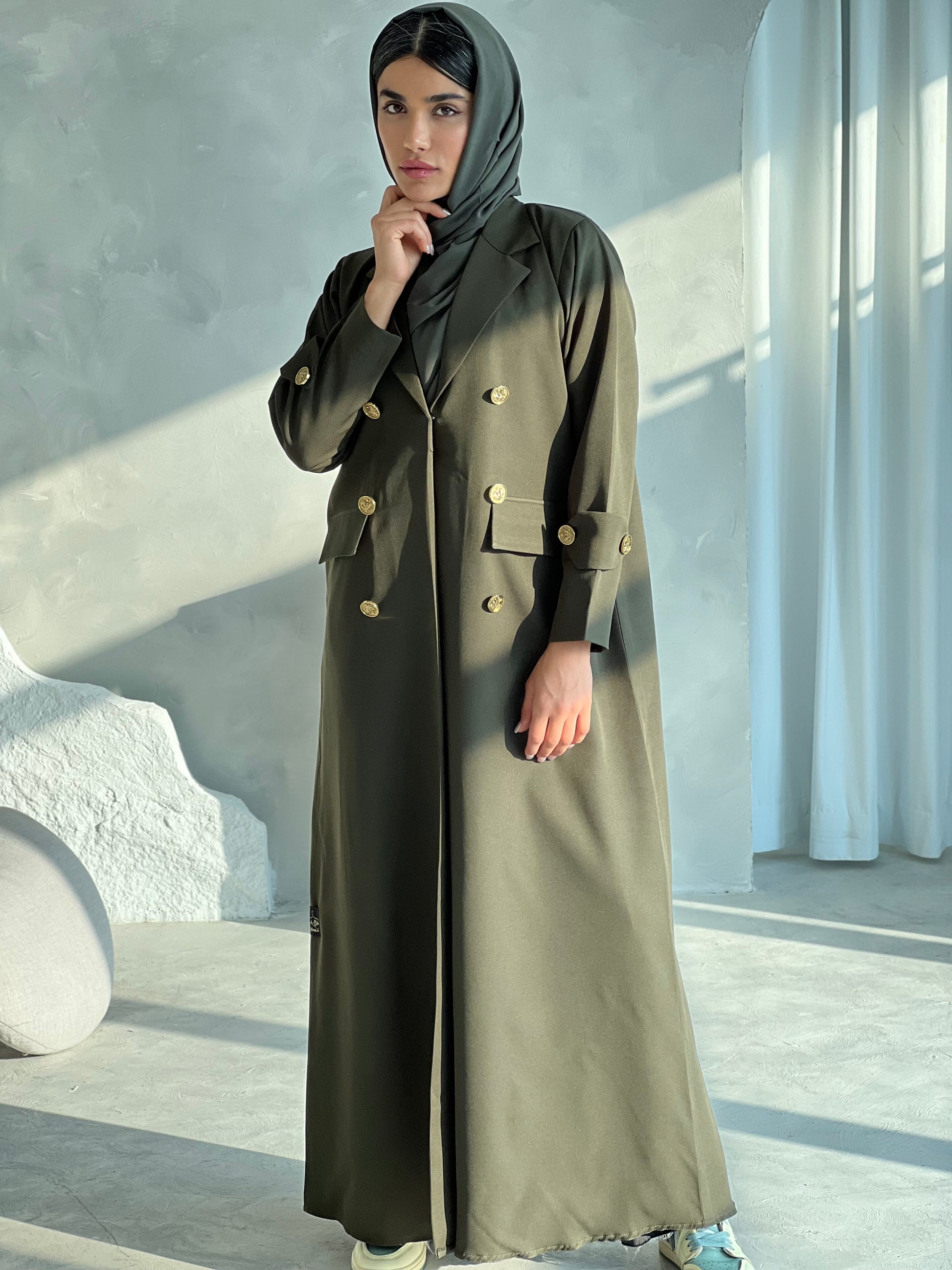 A woman wearing an Olive Green coat-style Abaya with a lapel collar and statement gold buttons with a matching Sheila