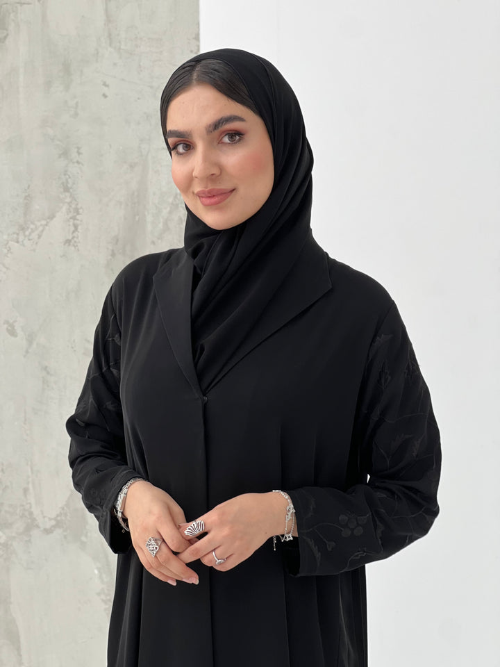 A woman wearing a Black Pleated Abaya with Floral Details, with long sleeves and a matching Sheila, in an indoor setting.