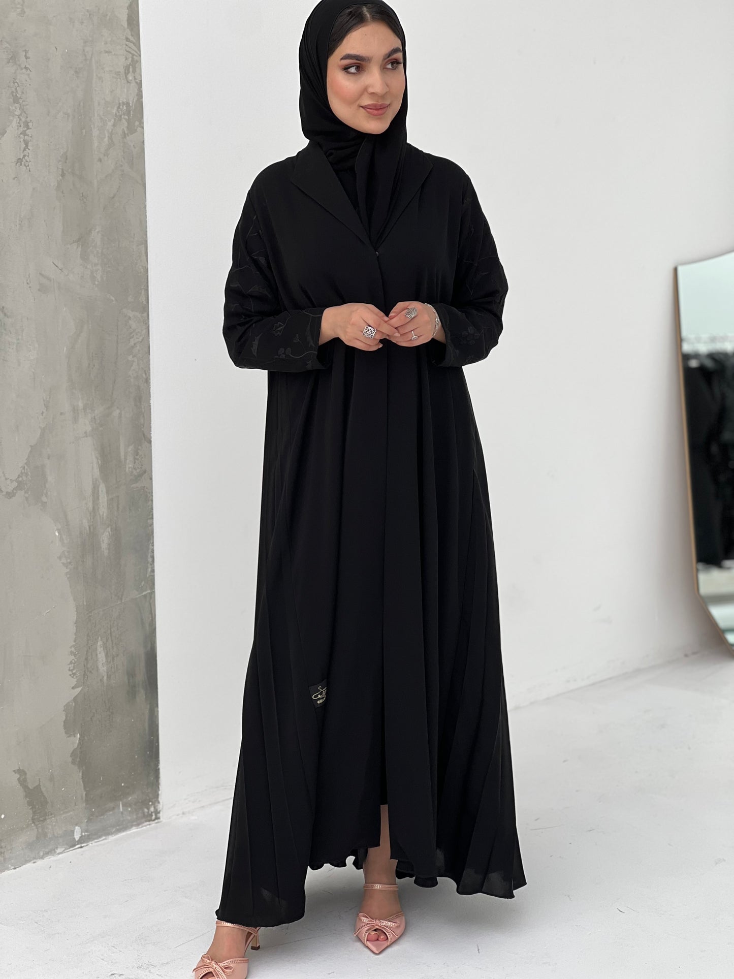 A woman wearing a Black Pleated Abaya with Floral Details, with long sleeves and a matching Sheila, in an indoor setting.