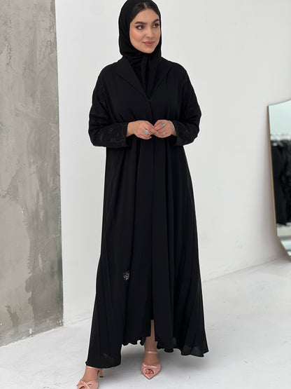 A woman wearing a Black Pleated Abaya with Floral Details, with long sleeves and a matching Sheila, in an indoor setting.