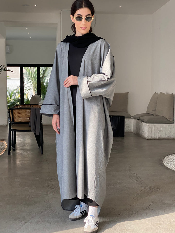 A woman wearing a Grey Cotton abaya with pants and a top, standing in a room with light decor.