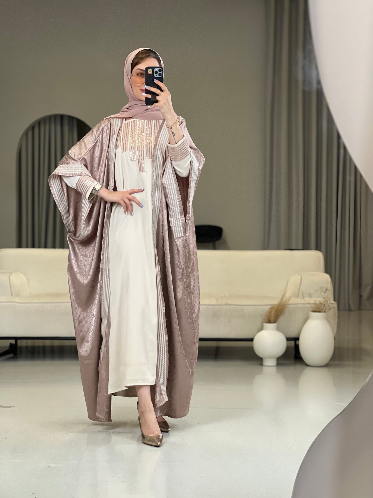 A  Woman wearing a Dusty Pink abaya with an embellished inner dress and a matching Sheila headscarf, standing in a room with minimalist decor