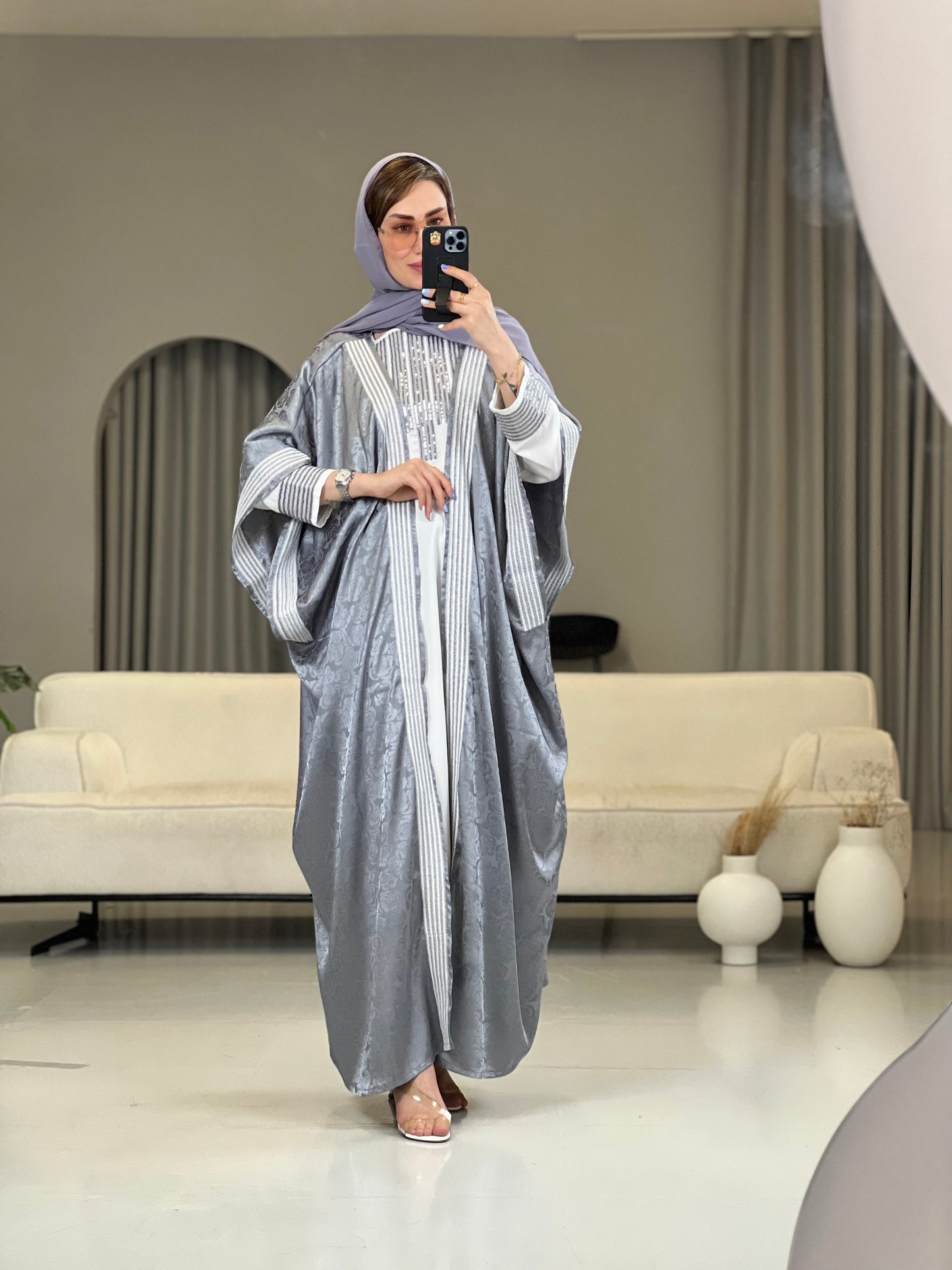 A  Woman wearing a Silver grey abaya with an embellished inner dress and a matching Sheila headscarf, standing in a room with minimalist decor