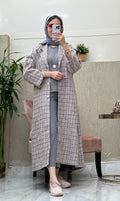 A woman wearing a Dusty Pink checkered tweed abaya with pants and a blouse, paired with a scarf and sneakers, standing in a room with a mirror and furniture.