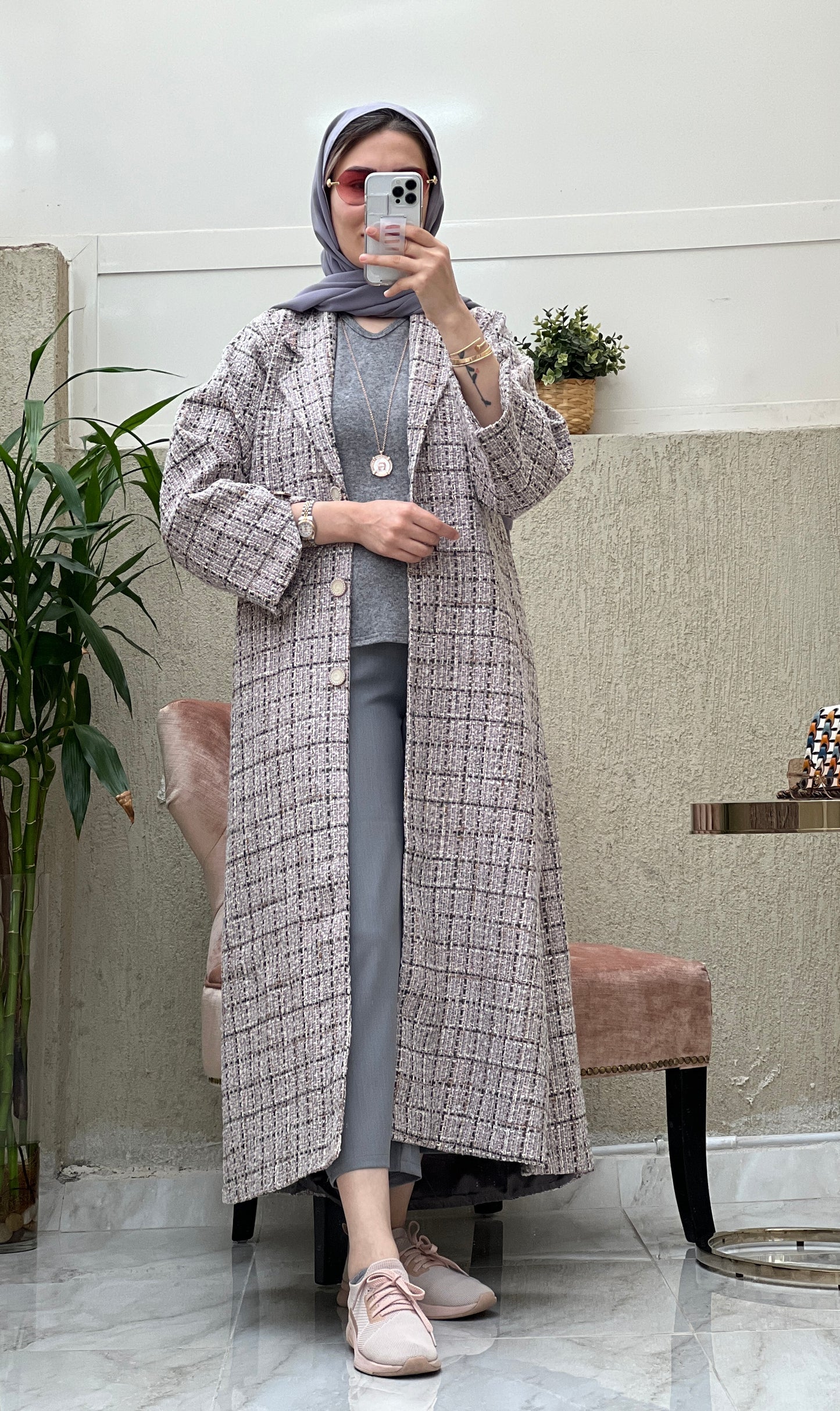 A woman wearing a Dusty Pink checkered tweed abaya with pants and a blouse, paired with a scarf and sneakers, standing in a room with a mirror and furniture.