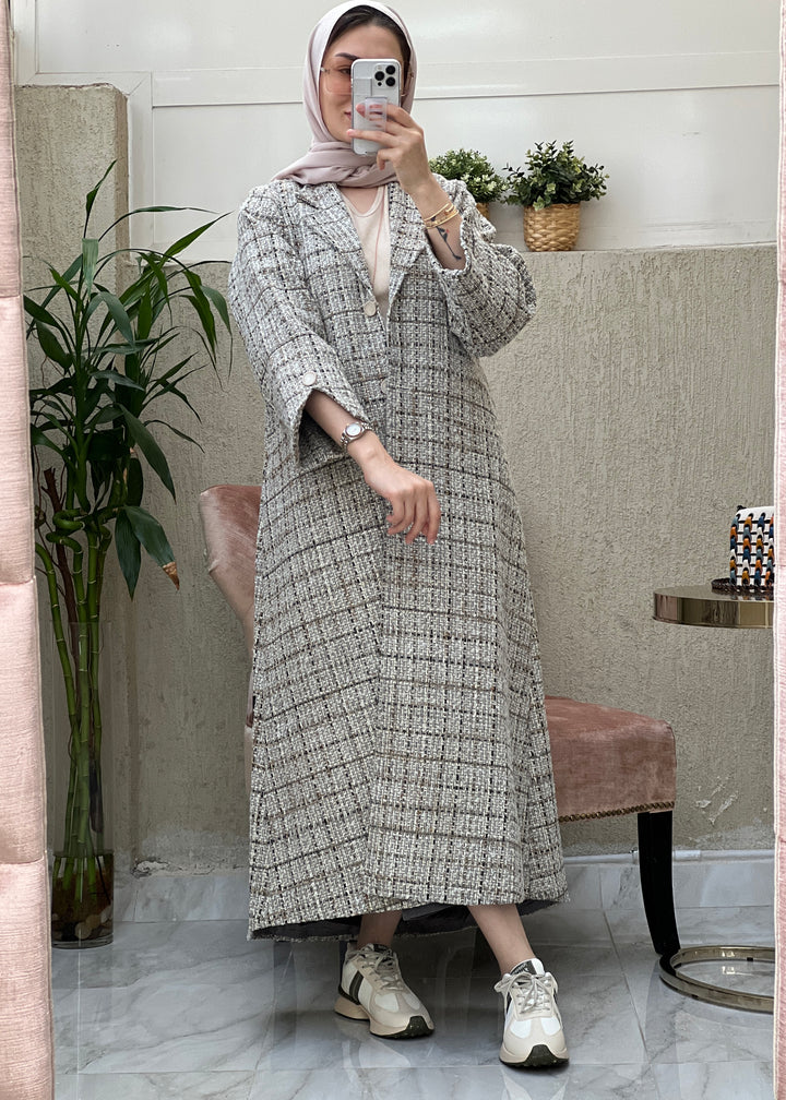 A woman wearing a grey checkered tweed abaya with pants and a blouse, paired with a scarf and sneakers, standing in a room with a mirror and furniture.
