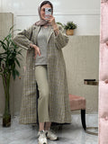 A woman wearing an Olive Green checkered tweed abaya with pants and a blouse, paired with a scarf and sneakers, standing in a room with a mirror and furniture.