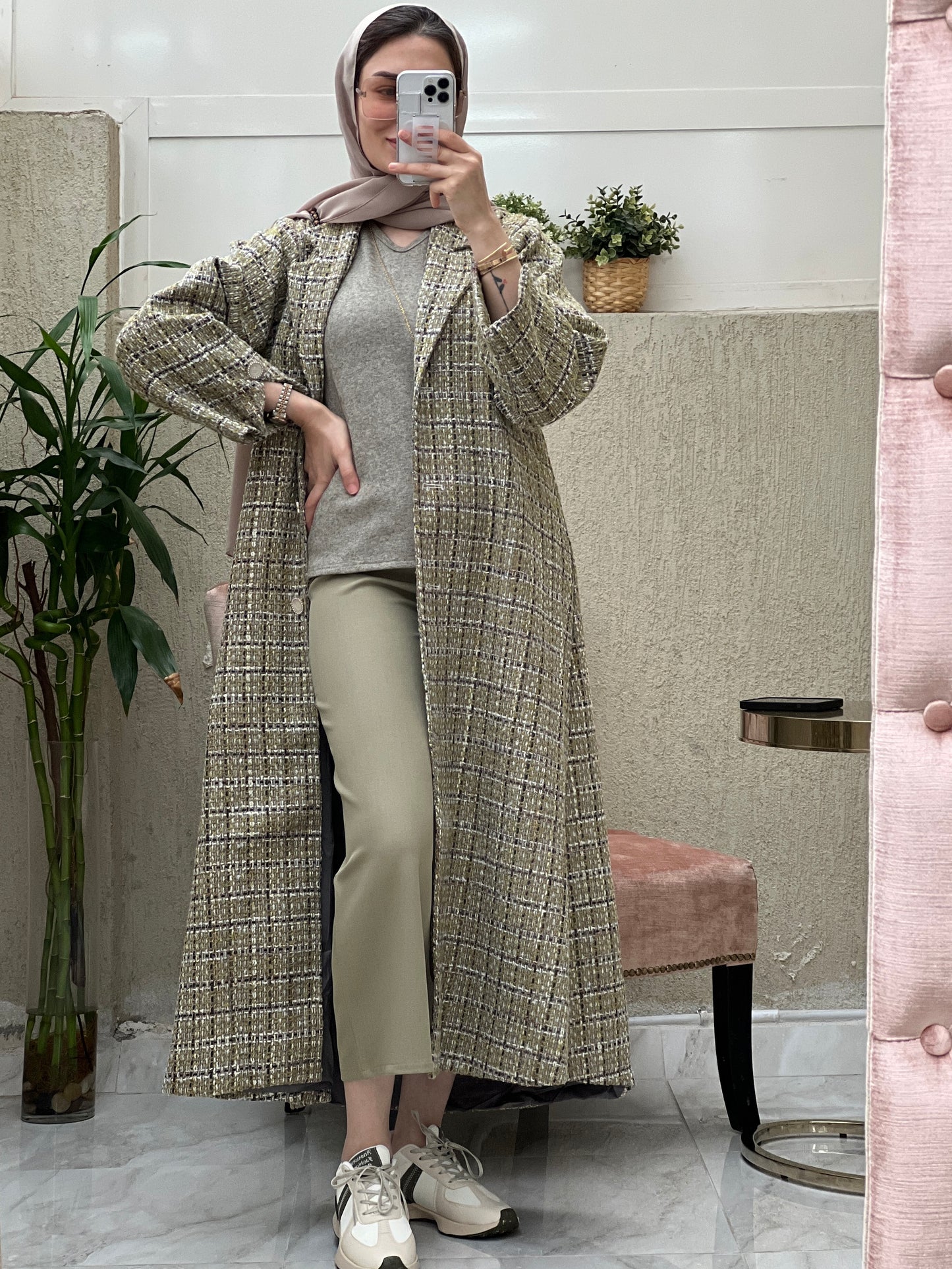 A woman wearing an Olive Green checkered tweed abaya with pants and a blouse, paired with a scarf and sneakers, standing in a room with a mirror and furniture.
