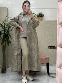 A woman wearing an Olive Green checkered tweed abaya with pants and a blouse, paired with a scarf and sneakers, standing in a room with a mirror and furniture.