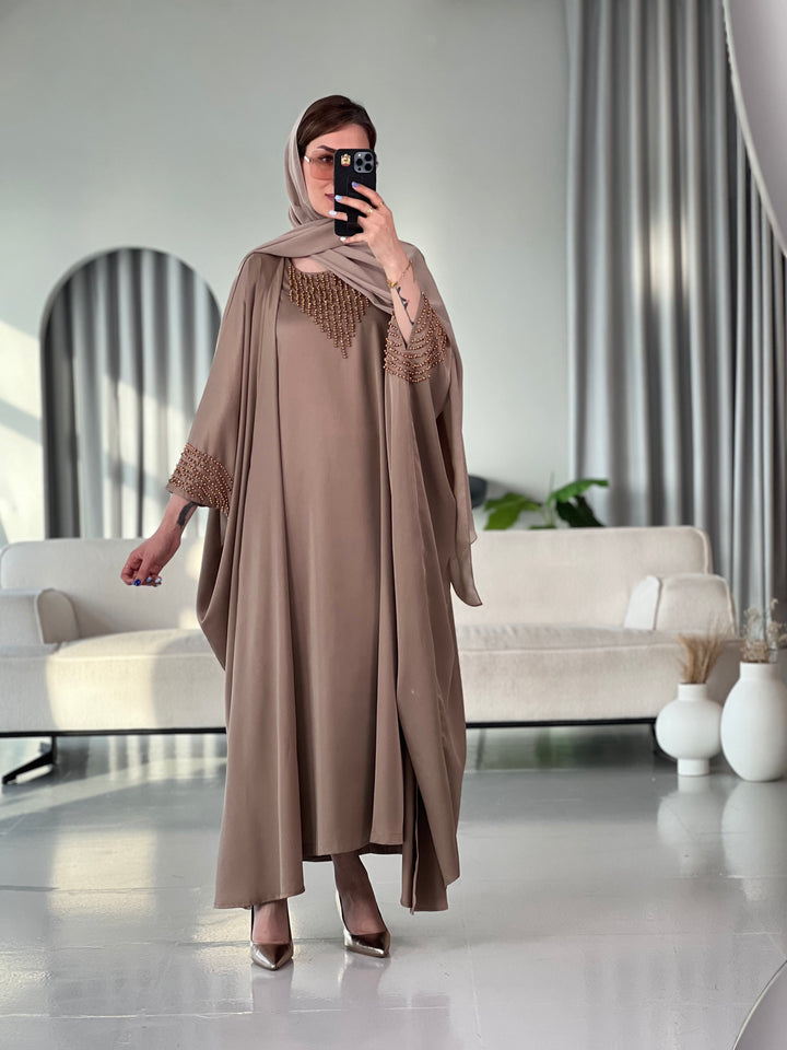 A woman standing in a room with white furniture, wearing a Light Brown / Mocha with Bronze Beading satin, Open front abaya style with embellished sleeves and inner and matching sheila