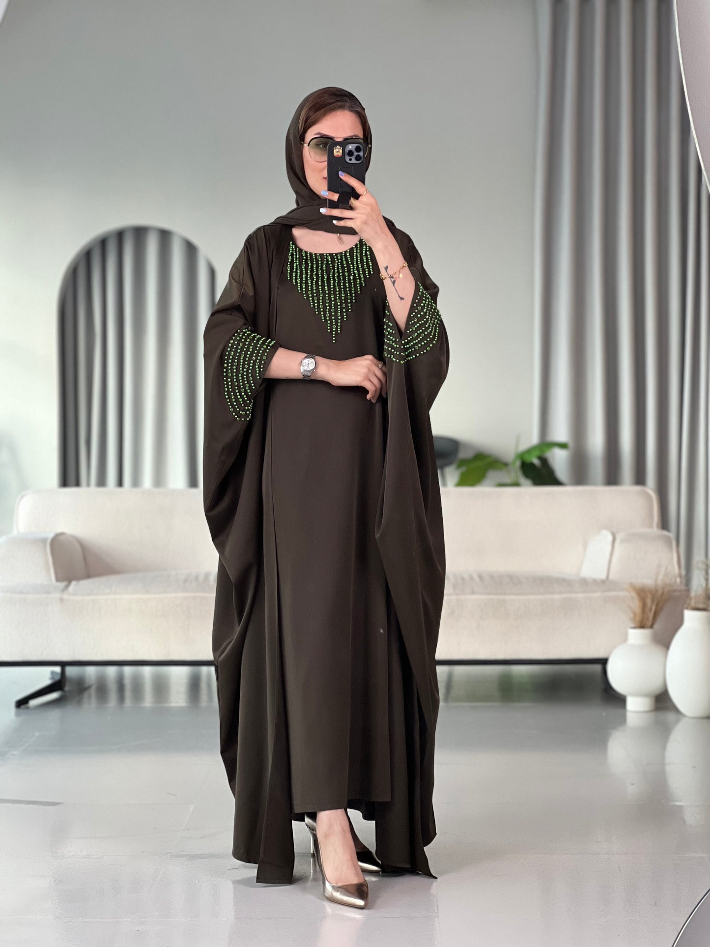 A woman standing in a room with white furniture, wearing a Dark Olive Green with Bright Green Beading satin, Open front abaya style with embellished sleeves and inner and matching sheila