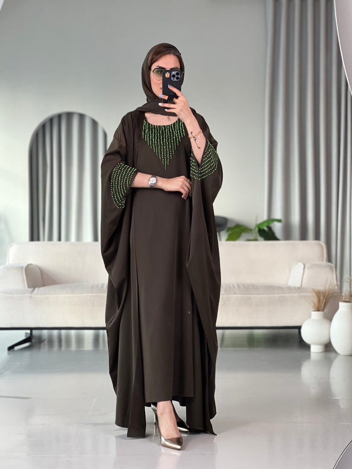 A woman standing in a room with white furniture, wearing a Dark Olive Green with Bright Green Beading satin, Open front abaya style with embellished sleeves and inner and matching sheila