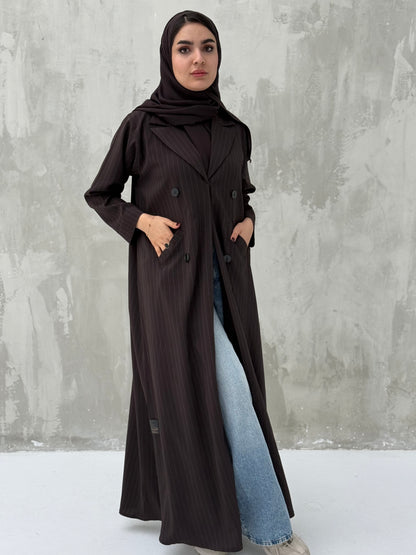 A woman wearing a brown abaya with a sheila, featuring a striped pattern, double-breasted design with buttons, and a V-neckline on a grey concrete background